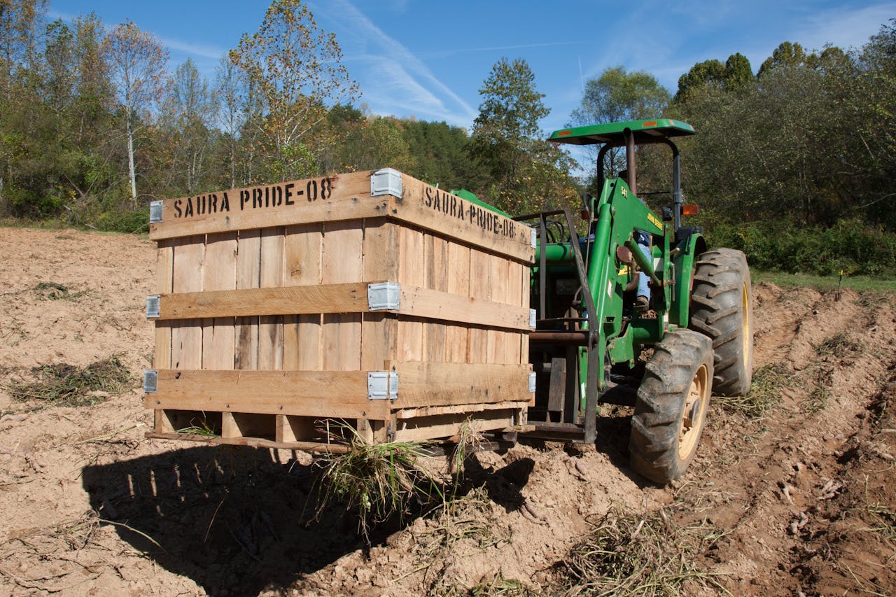 A green tractor in a field carrying a large wooden crate labeled Saura Pride 08 on a sunny day.
