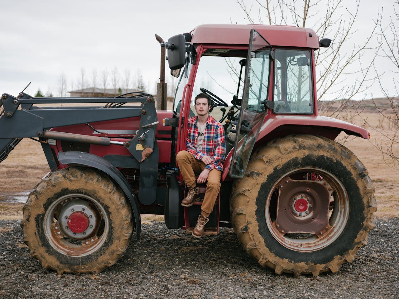 A young man in plaid shirt sits on a red tractor in a rural setting, showcasing rustic lifestyle.