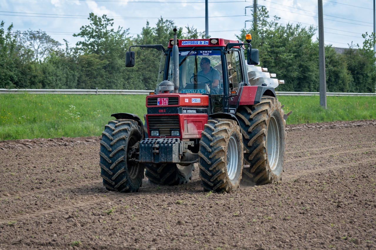 A red tractor is plowing a large field surrounded by greenery, preparing land for planting.